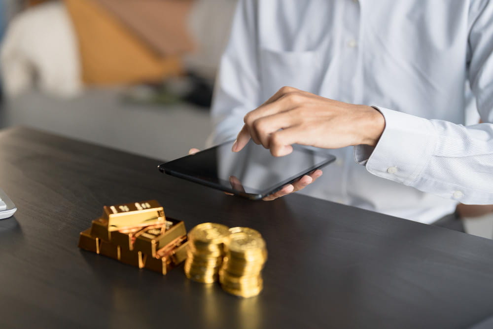 A professional using a digital tablet at a desk with gold bullion bars and stacked gold coins in the foreground, representing professional testing and finalizing your gold deal.